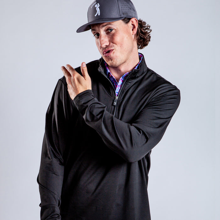 A person with long curly hair wearing a gray baseball cap and a Black - Q-Zip stands against a plain light background, pointing sideways and making a playful, exaggerated facial expression.