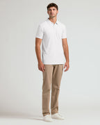A man modeling a white short-sleeve polo shirt, paired with beige trousers and white sneakers, standing against a light gray studio background.