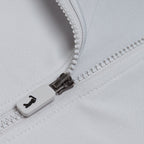 Close-up of the Bone - Q-Zip's white plastic zipper, partially unzipped on light gray, moisture-wicking fabric. The zipper pull displays a small black icon resembling a stylized seated figure.