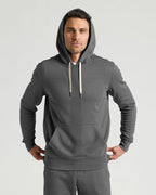 A man wearing a dark gray pullover hoodie with light beige drawstrings and matching gray sweatpants, standing against a light gray studio background.