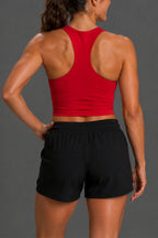 Rear view of a woman wearing a bright red racerback crop top and black athletic shorts against a dark gray studio background. || Black