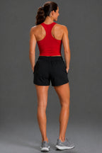 A woman shown from behind wearing a red athletic tank top and black athletic shorts, paired with gray sneakers, against a solid gray studio background. || Black