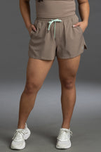 A model wearing taupe-brown athletic shorts with a mint green drawstring, paired with white sneakers, shown from the waist down against a gray studio background. || Mocha