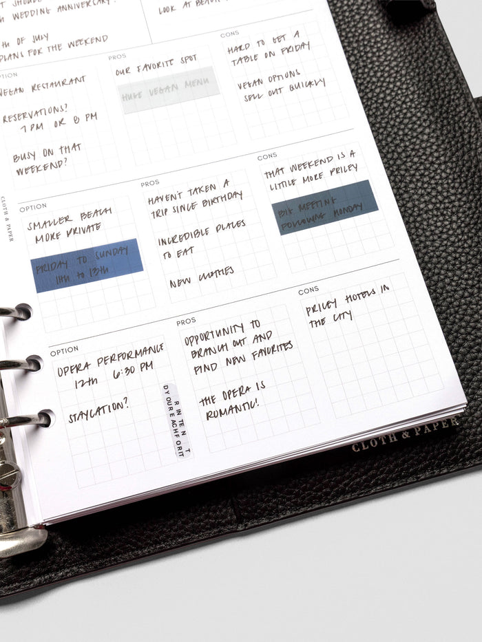 Close-up of an A5 decision-making insert inside a black leather planner, filled with handwritten options, pros, and cons in the gridded sections.