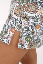 A model wearing a white A-line mini skirt with a colorful floral and botanical print, paired with white sneakers, shown against a plain light background.