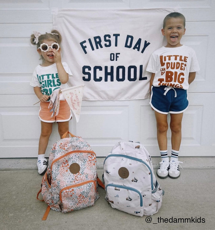 Two children standing in front of a 'First Day of School' sign with backpacks.