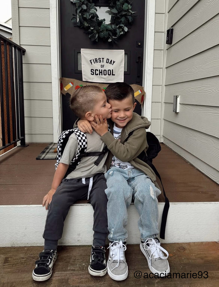 Two children sitting on a step outside a house with a 'First Day of School' sign.