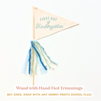 a small, beige-colored triangular flag with the text "FIRST DAY of Kindergarten" printed on it. The flag has a blue and white tassel attached to it.