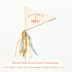 a hand-tied trimming on a white background, with a triangular flag attached to it that says "FIRST DAY OF 1ST GRADE".