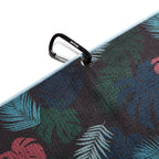 A close-up of the Haolie Nights - Towel shows its dark, tropical pattern with green, blue, and red leaves. A black carabiner clips to a metal eyelet at the light blue-edged corner. Enjoy vibrant 2-sided print design.