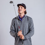 A man in a dark baseball cap and Heather Gray - Q-Zip holds a golf club upright, smirking as he looks up. Against a light gray background, his colorful patterned shirt peeks out beneath the athletic fit quarter-zip sweater.