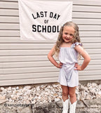 Young girl in a light blue dress standing in front of a 'Last Day of School' sign.