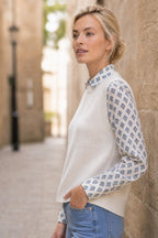 Model wearing the Aspiga Merino Wool Knitted Short Tank in Ivory layered over a printed blouse with jeans, side view leaning against a stone wall