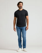 A man wearing a black Pima Cotton Blend Curved Hem Crew 4-Pack shirt paired with blue jeans and white sneakers, standing against a plain light background.