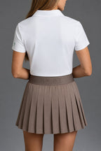 Back view of a woman wearing a white short-sleeve polo shirt and a pleated taupe-brown skirt against a solid gray studio background. || Mocha