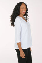 A woman with long curly black hair wearing a white textured three-quarter sleeve top and black pants, posing against a plain white studio background.