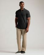 A man modeling a black short-sleeve polo shirt paired with beige trousers and black shoes, standing against a light gray studio background.