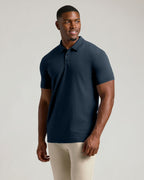 A man wearing a short-sleeve polo shirt in dark navy blue, paired with light beige pants, standing against a solid light gray studio background.